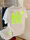 A white short-sleeve T-shirt hanging on a clear hanger, featuring a large neon yellow-green 3D block letter 'Hi' on the back with a matching neon yellow-green dot above it. A circular inset shows a close-up of the neon graphic on the front chest with a gold necklace worn over it.
