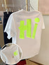 A white short-sleeve T-shirt hanging on a clear hanger, featuring a large neon yellow-green 3D block letter 'Hi' on the back with a matching neon yellow-green dot above it. A circular inset shows a close-up of the neon graphic on the front chest with a gold necklace worn over it.