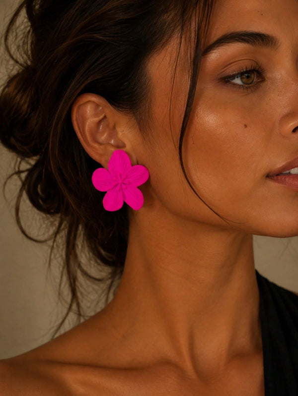 A close-up of a woman wearing a bright hot pink flower-shaped dangle earring. The earring has five layered petals and is shown on her earlobe against a warm, neutral background.