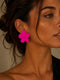 A close-up of a woman wearing a bright hot pink flower-shaped dangle earring. The earring has five layered petals and is shown on her earlobe against a warm, neutral background.