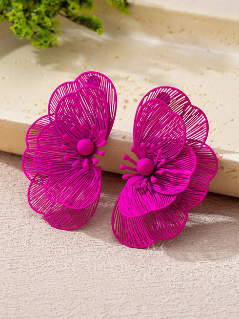 A pair of bright magenta-pink flower-shaped earrings with intricate wireframe petal detailing and small central spheres, displayed on a textured beige surface with a light stone slab in the background.