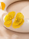 A pair of bright yellow flower-shaped earrings with fine textured petals and yellow centers, displayed on a curved cream-colored surface.