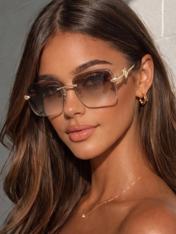 A close-up of a woman wearing oversized square sunglasses with a gold-tone metal frame and gradient brown lenses. She is also wearing small gold hoop earrings and a delicate gold necklace, set against a neutral gray background.