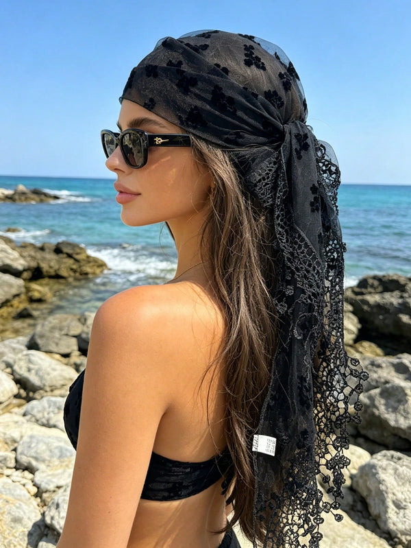 A woman on a rocky beach wearing a sheer black headscarf with floral embroidery and a long lace-trimmed tail, along with dark sunglasses.