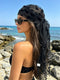 A woman on a rocky beach wearing a sheer black headscarf with floral embroidery and a long lace-trimmed tail, along with dark sunglasses.