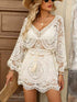Woman wearing a white lace dress with long sleeves and a hat, standing against a neutral background.