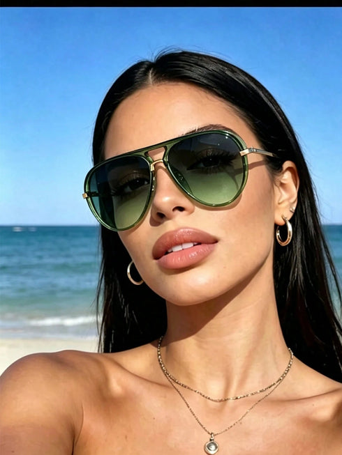 A woman on a beach wearing aviator sunglasses with olive green lenses and gold-toned metal frames, along with gold hoop earrings and layered necklaces.