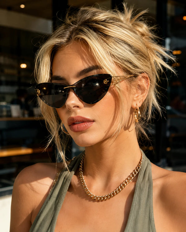 A close-up lifestyle shot of a woman with blonde hair wearing stylish cat-eye sunglasses with gold frames, a gold chain necklace, and a green halter top.