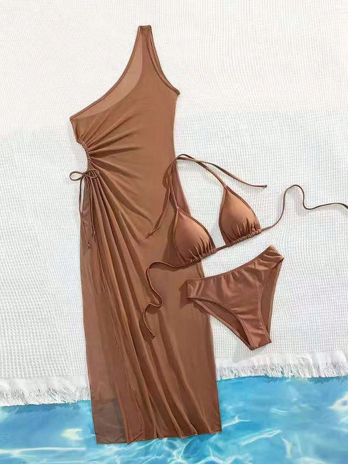A brown swimwear set laid flat on a white textured surface with a blue water-patterned strip at the bottom. The set includes a one-shoulder maxi cover-up dress with side ties, a triangle bikini top, and matching bikini bottoms.