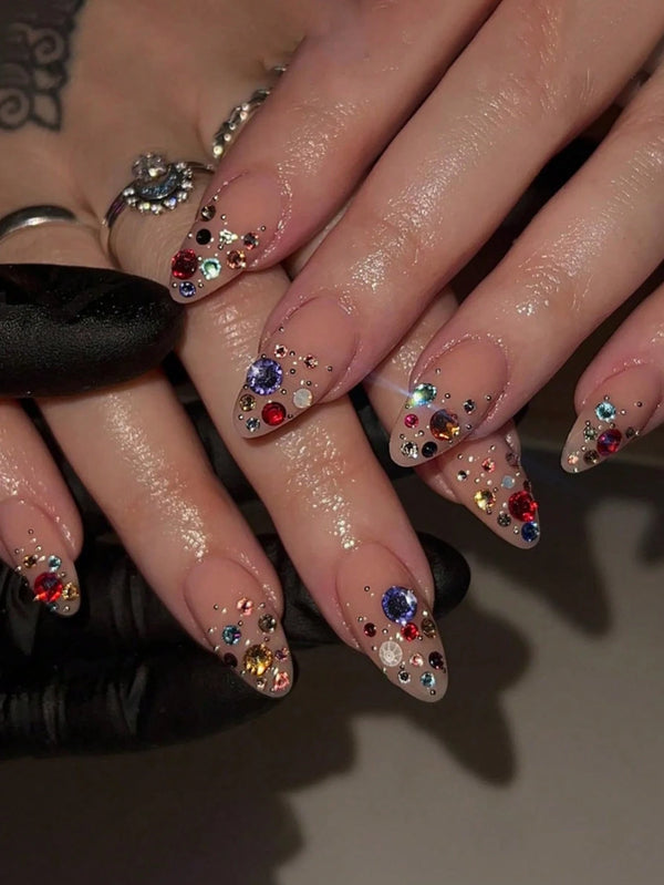 Close-up of manicured fingernails with glossy nude bases, glitter accents, and colorful rhinestone clusters, shown on a hand with rings and a black glove.