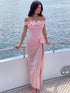 A woman models an off-the-shoulder pink maxi dress with 3D floral details on the neckline and a matching flower at the waist, standing on a yacht deck with the ocean in the background.