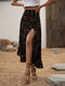 A woman wearing a black high-waisted midi skirt with a red and white floral print, paired with a white crop top and brown strappy high-heeled sandals, standing in an outdoor rustic setting with sand and stones.