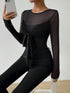 A woman wearing a black sheer long-sleeve top with a twisted front detail and black skinny pants, holding a phone in a mirror selfie against a textured white wall.