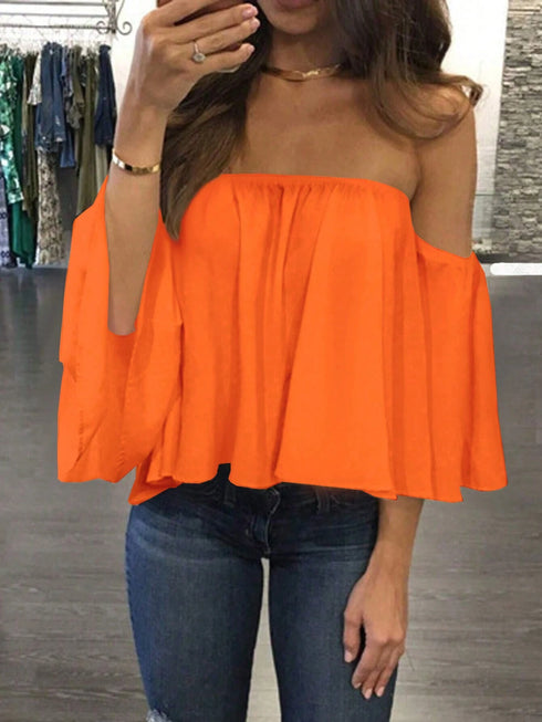 A woman wearing a bright orange off-the-shoulder top with wide bell sleeves and blue jeans, taking a mirror selfie in a clothing store.