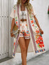 A woman models a bohemian-style two-piece outfit featuring a cream-colored short-sleeve open-front kimono jacket and matching crop shorts, both decorated with vibrant red, orange, and green floral prints and colorful geometric borders. She is also wearing a white crop top and holding a woven clutch.