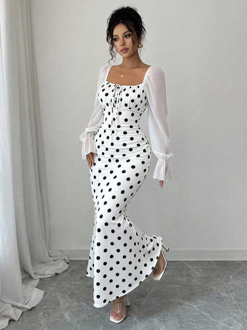 A woman models a white maxi dress with black polka dots and sheer white long sleeves, standing in a minimalist room with a light gray wall and floor.
