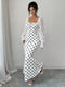 A woman models a white maxi dress with black polka dots and sheer white long sleeves, standing in a minimalist room with a light gray wall and floor.