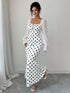 A woman models a white maxi dress with black polka dots and sheer white long sleeves, standing in a minimalist room with a light gray wall and floor.