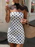 A woman wearing a white strapless mini dress with black polka dots and a black lace mock neck, holding an ice cream cone and a straw bag in an outdoor setting.