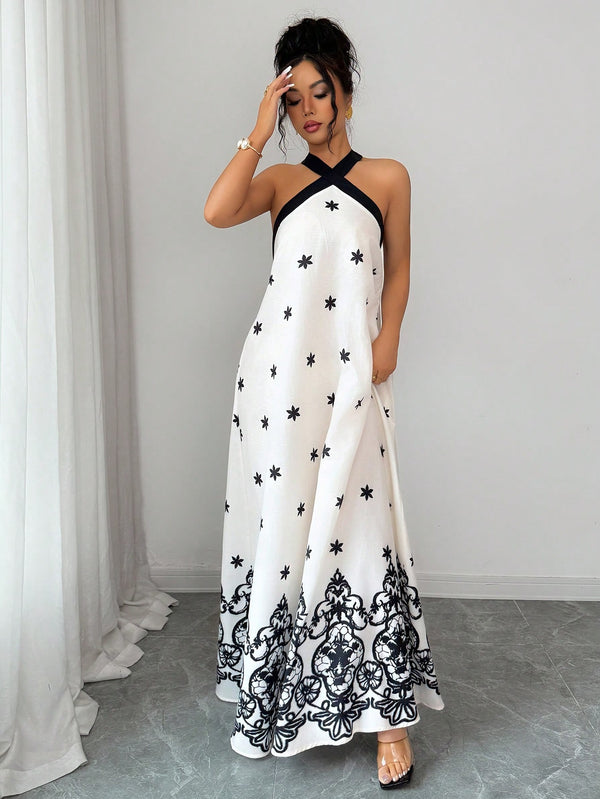 A woman models a white halter maxi dress with black star and damask patterns, standing in a minimalist room with a gray floor and white curtains.