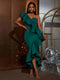 A woman models a dark green one-shoulder ruffled gown with a high-low hem, paired with gold strappy heels and hoop earrings, standing in a decorated indoor setting with disco balls and a gold frame.