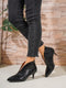 Black crocodile-patterned high-heeled shoes worn with black jeans against a wooden wall.