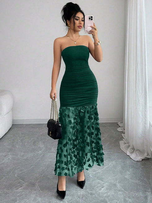 A woman takes a mirror selfie wearing a strapless dark green ruched maxi dress with a sheer floral patterned lower skirt. She is holding a black quilted handbag and wearing black pointed-toe heels in a bright room with gray tiled floors and white curtains.