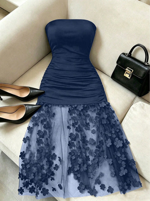 A navy blue strapless dress with a ruched bodice and a sheer tulle skirt adorned with 3D floral appliqués, styled on a beige sofa alongside black pointed shoes and a black handbag.