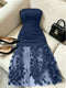 A navy blue strapless dress with a ruched bodice and a sheer tulle skirt adorned with 3D floral appliqués, styled on a beige sofa alongside black pointed shoes and a black handbag.