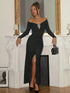 A woman models a long-sleeve black off-the-shoulder maxi dress with a thigh-high slit and a silver geometric accent at the neckline, standing in front of a white fireplace mantel.