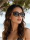 A woman with long wavy brown hair wearing black cat-eye sunglasses with gold metal accents, photographed outdoors near a beach with trees in the background.