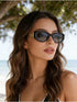 A woman with long wavy brown hair wearing black cat-eye sunglasses with gold metal accents, photographed outdoors near a beach with trees in the background.