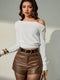 A woman modeling a white ribbed off-the-shoulder long-sleeve crop top paired with brown faux-leather shorts and a black belt, set against a neutral studio background.