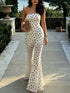 A woman wearing a strapless white maxi dress with black polka dots, standing on a stone pathway in an outdoor garden setting.