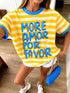 A woman wearing a yellow and white striped ringer t-shirt with blue trim and the text 'MORE AMOR POR FAVOR' in blue letters, paired with white shorts and holding a blue purse.