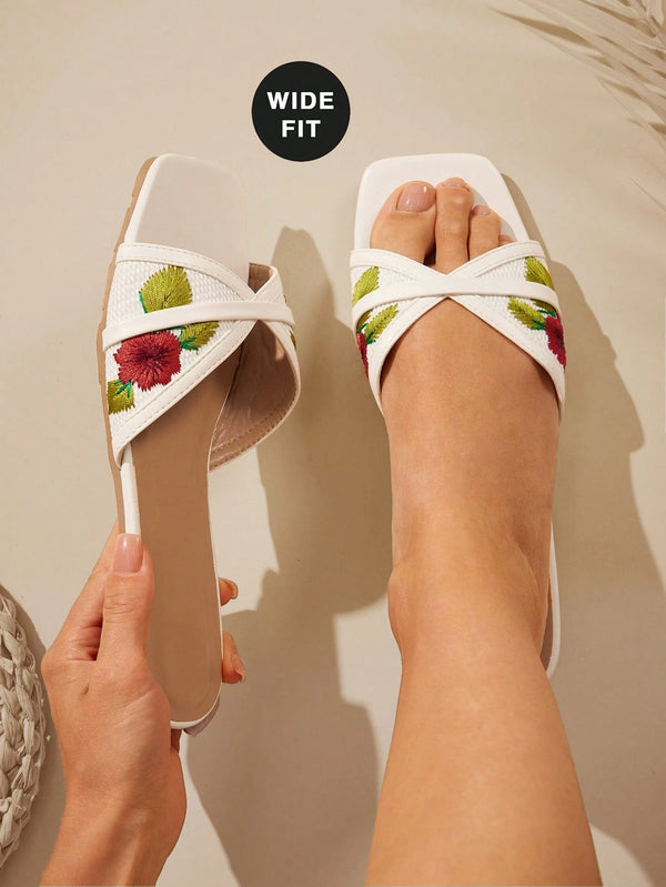 White wide-fit slide sandals with colorful floral embroidery, shown with one sandal worn on a foot and the other held by a hand against a neutral beige background.