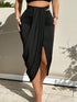 A woman wearing a black high-waisted midi skirt with ruched detailing, side cutouts, and a front slit, standing outdoors against a beige wall with leaf shadows.