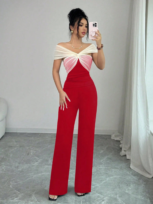A woman takes a mirror selfie wearing a red wide-leg jumpsuit with a cream and pink twisted off-the-shoulder bodice, standing in a room with gray tiled floors and white curtains.