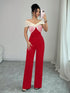 A woman takes a mirror selfie wearing a red wide-leg jumpsuit with a cream and pink twisted off-the-shoulder bodice, standing in a room with gray tiled floors and white curtains.