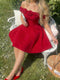 A woman wearing a bright red off-the-shoulder mini dress with 3D rose details on the neckline, sitting on a white garden bench outdoors.