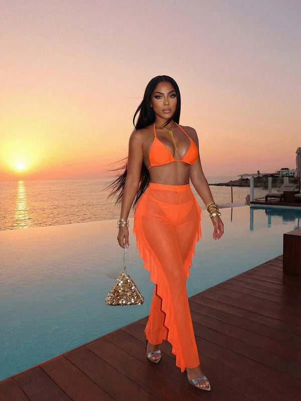 A woman stands on a wooden deck beside an infinity pool at sunset, wearing a bright orange two-piece outfit with a halter top and a long sheer fringe skirt. She is holding a small glittering triangle handbag.