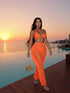 A woman stands on a wooden deck beside an infinity pool at sunset, wearing a bright orange two-piece outfit with a halter top and a long sheer fringe skirt. She is holding a small glittering triangle handbag.