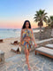 A woman wearing a colorful, patterned two-piece swimwear set with a halter top and matching mini skirt, walking on a stone path at a beach resort during sunset.