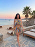 A woman wearing a colorful, patterned two-piece swimwear set with a halter top and matching mini skirt, walking on a stone path at a beach resort during sunset.