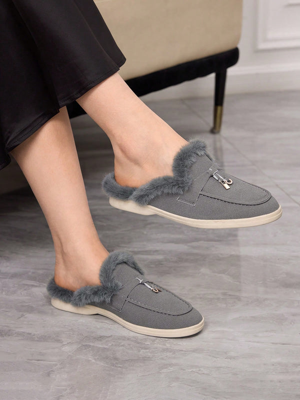 Gray slip-on slippers with fluffy faux-fur trim and a small silver zipper pull, shown on a person's feet in a home setting with a gray tile floor and a beige chair visible.
