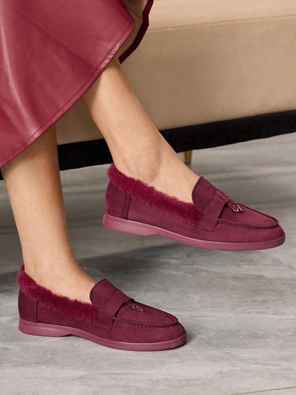 A person wearing burgundy suede slip-on loafers with plush faux-fur trim and a matching sole, shown in a styled indoor setting with a sofa in the background.