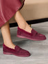 A person wearing burgundy suede slip-on loafers with plush faux-fur trim and a matching sole, shown in a styled indoor setting with a sofa in the background.