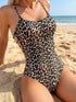 A woman wearing a leopard print one-piece swimsuit sits on a sandy beach with the ocean and blue sky in the background.