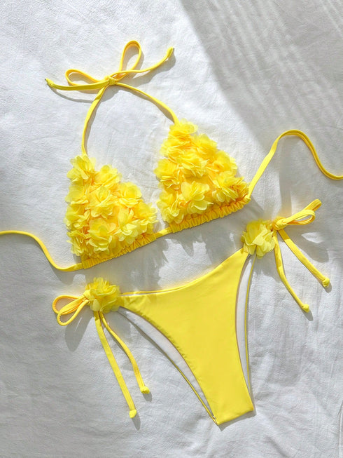 A bright yellow two-piece bikini set laid flat on a white fabric background. The triangle bikini top has textured, ruffled flower-like detailing on each cup and thin side ties.