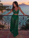 A woman stands on a tropical beach balcony at sunset wearing a green two-piece cutout lace outfit with a halter crop top and matching wide-leg skirt. The background shows ocean water, palm trees, and distant mountains.